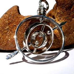 May include: A silver pendant with a circular design featuring a smaller circle and a spiral inside. The pendant is suspended from a silver chain and has a small arrow-shaped piece extending from the side.