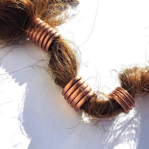 Puede incluir: Primer plano de cabello castaño adornado con cuentas para el cabello de color cobre. Las cuentas son cilíndricas con detalles acanalados, agregando textura. El cabello es de un color marrón cálido, y las cuentas parecen ser de metal.