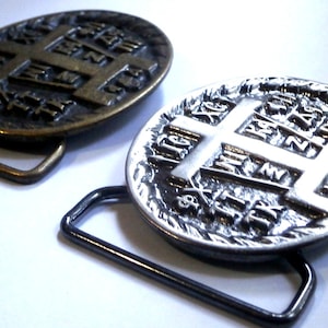 May include: Two oval belt buckles, one in a dark bronze finish and one in a bright silver finish. Each buckle features a central cross design with embossed Greek lettering and symbols, and a rectangular metal loop attached to the base for a belt.
