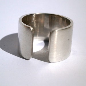 Könnte beinhalten: Ein silberner Ring mit einem breiten Band und einer Lücke in der Mitte. Der Ring ist aus glattem Metall gefertigt und hat eine polierte Oberfläche.