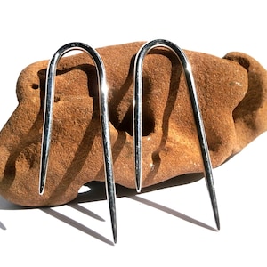 May include: A pair of silver-colored, U-shaped hair pins. The pins are resting on a rough, brown stone against a white background. The pins have a polished, reflective surface and a pointed end.