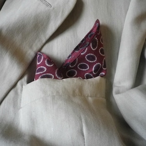 May include: Close-up of a light beige blazer with a burgundy pocket square. The pocket square features a repeating pattern of white ovals on a burgundy background. The blazer's pocket is visible.