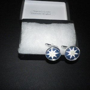 May include: A pair of silver cufflinks with a blue and white star-shaped design. The cufflinks are in a black box with white tissue paper. The box has a label that reads "Yates & Co London Hand made in England."
