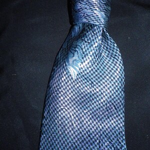 May include: A blue necktie with a checkered pattern and a floral design. The tie is knotted, showing its texture and the interplay of light and shadow. The background is a solid black.