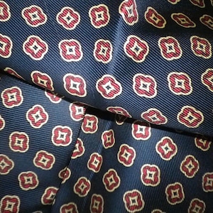 May include: Close-up of navy blue fabric with a repeating pattern of red and white floral shapes. The fabric is folded, showing the texture and design. The pattern is evenly spaced.