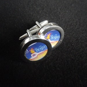 以下が含まれることがあります： ビートルズのロゴとギターのイラストが描かれた円形デザインのシルバーカフスボタン。カフスボタンは光沢のある金属仕上げです。