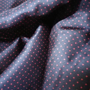 May include: Close-up of a navy blue fabric with a repeating pattern of small, evenly spaced red polka dots. The fabric is folded, creating shadows and highlighting the texture and sheen of the material.