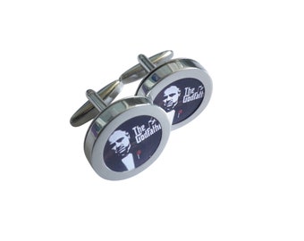 May include: A pair of silver cufflinks with a black and white image of a man's face and the text "The Godfather" on each cufflink.