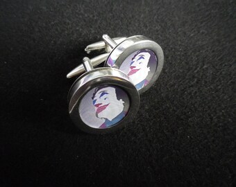 May include: A pair of silver cufflinks with a round, white background and a colorful image of a person's face. The person has blue eyes, pink lips, and a purple and blue patterned shirt.
