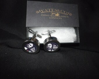 May include: A pair of silver cufflinks with a circular design featuring a portrait and the text "The Godfather" on a dark blue background. The cufflinks are presented in a black box with the text "AYATES & CO HAND MADE LONDON".