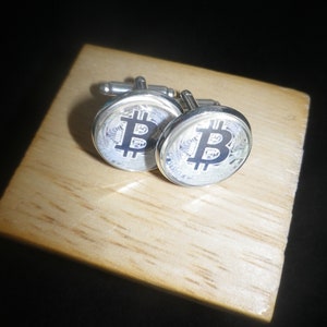 May include: A pair of silver cufflinks with a black and white Bitcoin logo on each cufflink. The cufflinks are on a wooden surface.