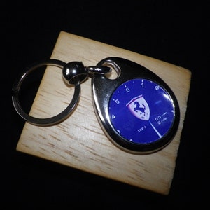 Peut inclure: Porte-clés en métal argenté avec un graphique bleu et blanc. Le porte-clés présente un fond bleu avec un emblème blanc et rouge et un design de compteur de vitesse. Le porte-clés est sur un bloc de bois.