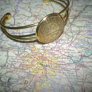 May include: A gold-toned bracelet with a round coin charm featuring the text "Liberté, Fraternité, 20 Centimes 1900". The bracelet is lying on a map of France.