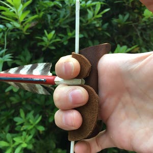 May include: A brown leather archery finger tab with a white string attached to it. The tab is worn on the index finger of a hand holding a bow and arrow.