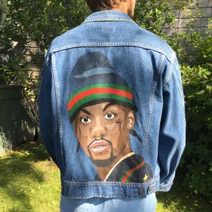 Puede incluir: Una chaqueta vaquera azul con un retrato pintado de una persona que lleva un sombrero negro y verde. La persona tiene piel morena, ojos marrones y bigote.