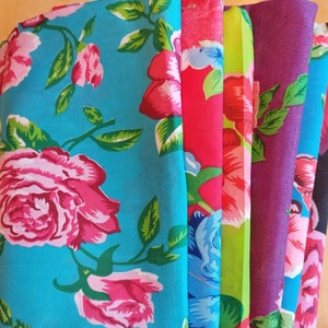 Può includere: Una pila di tessuti floreali colorati. I tessuti presentano grandi motivi di rose nei toni del rosa, rosso, blu e viola, con foglie verdi. I tessuti sono piegati e impilati su una superficie di legno.