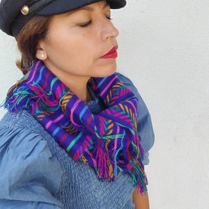 May include: A woman wearing a black newsboy cap with a braided band and a colorful fringed scarf. The scarf has a geometric pattern in shades of purple, pink, green, and yellow.