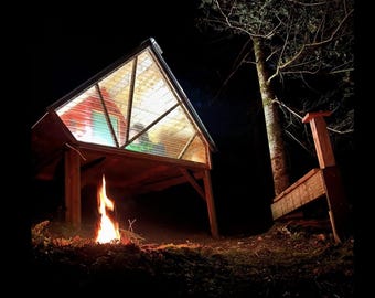 May include: A wooden cabin with a clear plastic roof and a fire burning in front of it. The cabin is lit up from the inside and there is a tree in the background.