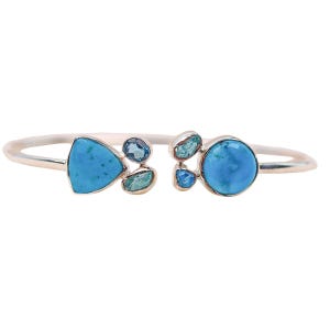 Op de afbeelding: Een zilverkleurige manchetarmband met turquoise en blauwe edelstenen. De armband heeft een driehoekige turkooissteen en een ronde turkooissteen, met clusters van kleinere blauwe en groene stenen.