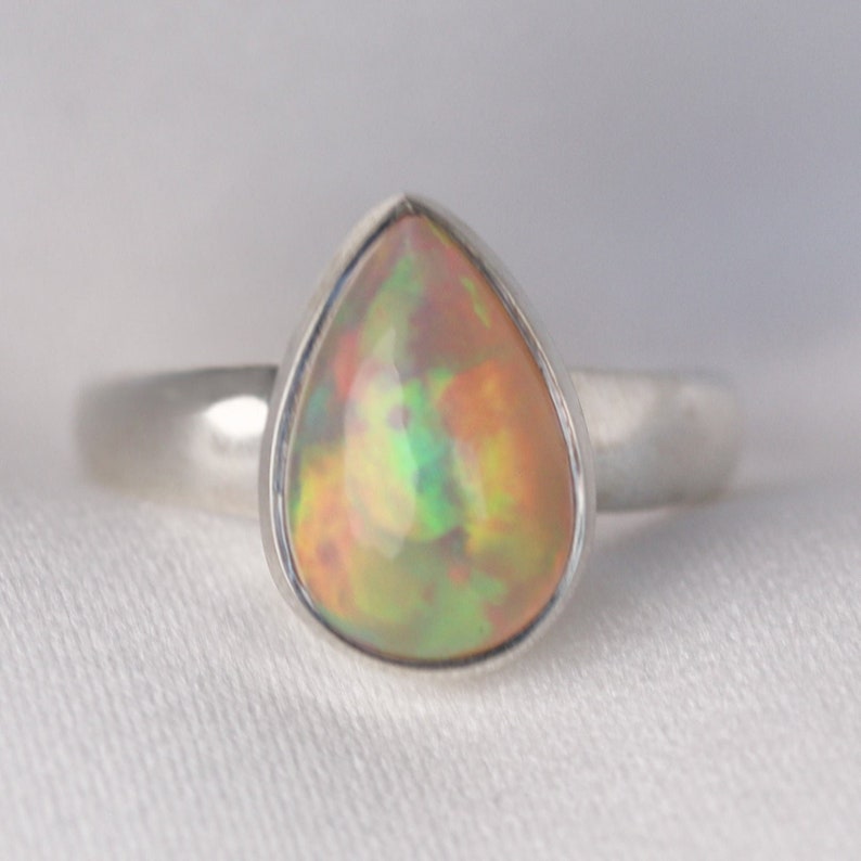 Puede incluir: Un anillo de plata con una opala en forma de l&aacute;grima. La opala es de color blanco lechoso con destellos de verde, naranja y amarillo.