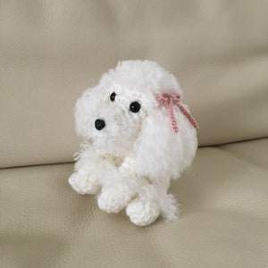 Peut inclure: Un jouet en crochet en forme de caniche blanc avec des yeux noirs et un n&oelig;ud rose. Le jouet est assis sur un canap&eacute; brun clair.