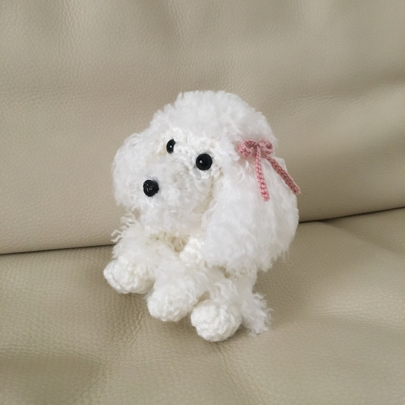 Peut inclure: Un jouet en crochet en forme de caniche blanc avec des yeux noirs et un n&oelig;ud rose. Le jouet est assis sur un canap&eacute; brun clair.