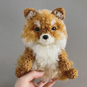 Puede incluir: Un peluche de zorro hecho a mano con ganchillo. El zorro tiene un aspecto esponjoso y texturizado con una mezcla de hilo marrón, blanco y beige. Tiene ojos negros y una nariz negra.