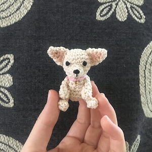 May include: A small crocheted chihuahua dog toy with a pink collar and a gold bell. The toy is being held in a person's hand.