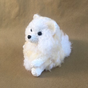 Puede incluir: Un animal de peluche blanco de crochet que se asemeja a un perro pomerania esponjoso. El perro está sentado con la cabeza girada hacia un lado y sus ojos son negros.