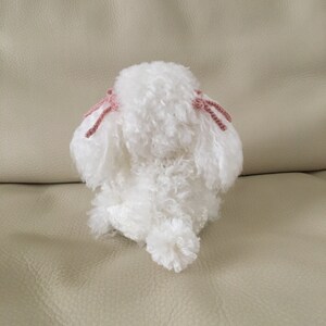 Peut inclure: Un chien en peluche blanc crochet&eacute; avec des n&oelig;uds en laine rose sur les oreilles. Le chien est assis sur un canap&eacute; brun clair.