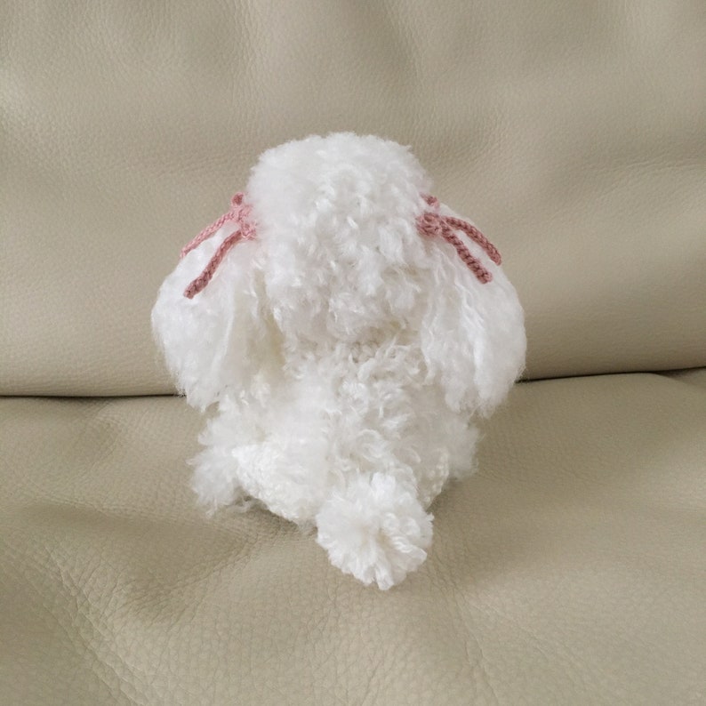 Peut inclure: Un chien en peluche blanc crochet&eacute; avec des n&oelig;uds en laine rose sur les oreilles. Le chien est assis sur un canap&eacute; brun clair.