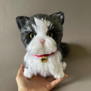 May include: A handmade plush cat with a black and white coat, green eyes, and a pink nose. The cat wears a red collar with a gold charm. The plush toy is soft and fluffy, and appears to be held in a hand.