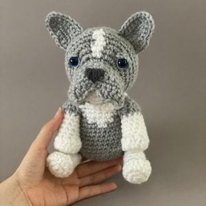 Puede incluir: Un juguete de peluche de bulldog francés de color gris y blanco tejido a crochet con ojos azules. El juguete está sentado en una mano.