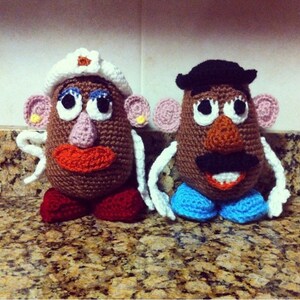 May include: Two crocheted plush toys of Mr. and Mrs. Potato Head.  Mr. Potato Head is wearing a black hat and a blue and white outfit. Mrs. Potato Head is wearing a white hat with a pink flower and a red and white outfit.