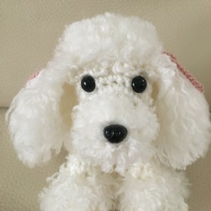 Peut inclure: Jouet en crochet en forme de caniche blanc avec des yeux noirs et un nez noir. Le caniche a des n&oelig;uds roses sur les oreilles.
