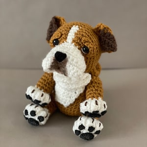 May include: A crocheted brown and white bulldog plush toy with black eyes and a black nose. The toy is sitting with its paws in front of it.