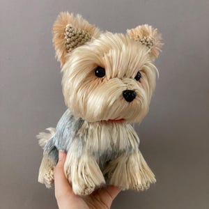 Può includere: Un peluche a forma di cane Yorkshire Terrier con lunghe fibre setose color beige e grigio. Il giocattolo presenta orecchie a punta lavorate all'uncinetto, occhi in plastica nera lucida, un piccolo naso nero e un sottile collare rosso.