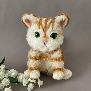 May include: A crocheted stuffed cat toy with white and orange fur, green eyes, and a pink nose. The cat is sitting on a gray background.