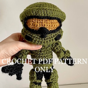 May include: A handmade crochet doll in the likeness of a video game character, primarily olive green with black and tan accents. The doll is holding a black crochet gun. Text on the image reads "CROCHET PDF PATTERN ONLY."