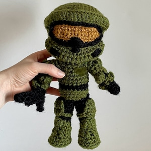 Puede incluir: Muñeco de ganchillo hecho a mano de un personaje con un traje verde y negro con una visera dorada. El muñeco está hecho de hilo y tiene un diseño detallado, que incluye un casco, una armadura y un arma. El muñeco mide aproximadamente 20 cm de altura.