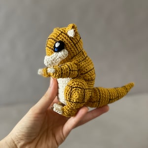 May include: A crocheted yellow and white amigurumi toy resembling a small dinosaur. The toy has black eyes and a long tail.