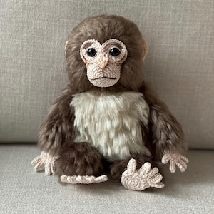 Op de afbeelding: Een handgemaakte pluchen aap met bruine vacht, een crèmekleurig gezicht en een witte borst. Het gezicht en de handen van de aap zijn gehaakt en hij heeft grote, donkere ogen. Het pluche speelgoed zit op een grijze ondergrond.
