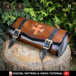 May include: A cylindrical brown leather tool bag featuring a centered tan cross emblem, black leather trim, and two black leather straps with brass-toned metal buckles. The bag has decorative scalloped edges and is designed for motorcycle storage.