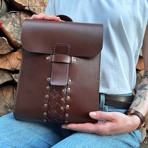 Puede incluir: Mochila de cuero marrón oscuro con cierre de solapa y detalle de cuero trenzado. La bolsa presenta remaches de latón y un cierre de hebilla. La mochila se sostiene frente a un fondo de madera apilada.