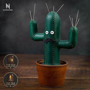 May include: A dark green leather cactus pincushion with visible stitching, two white button eyes, and a black leather mustache. The cactus is planted in a brown leather pot and features several silver sewing needles inserted into the top.