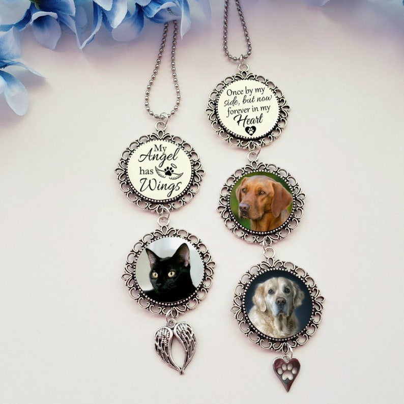May include: A silver car charm with three round ornaments. The top ornament has the text "Once by my side, but now forever in my Heart". The middle ornament has a photo of a brown dog. The bottom ornament has a photo of a golden retriever. The charm also has a silver angel wing and a silver paw print heart.