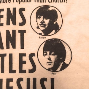 Może przedstawiać: Kremowa koszulka z czarnym tekstem i portretami Ringo i Paula z The Beatles. Tekst brzmi "ENS WANT BEATLES JESUS!" nad portretami. Portrety są w okrągłych ramkach.