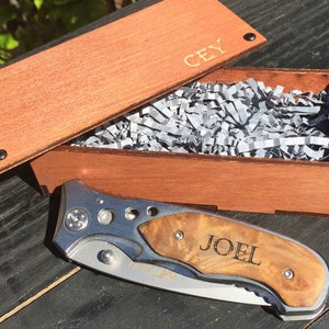 May include: A wooden box with a personalised inscription "CEY" on the lid. The box is open and contains a small knife with a wooden handle. The knife has the name "JOEL" engraved on the handle.