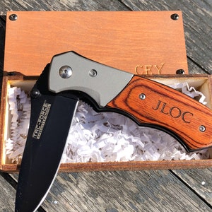 May include: A black folding knife with a wood handle engraved with the name "JLOC". The knife is in a wooden box with brown paper filler.