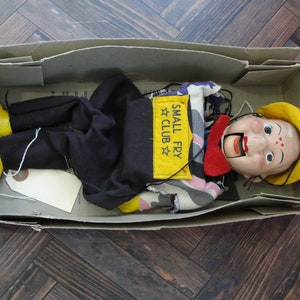May include: A vintage hand puppet in a box. The puppet is wearing a yellow hat, a red bow tie, and a black and white striped shirt. The puppet has a painted face with a big smile and a red mouth. The box is labeled "Small Fry Club".